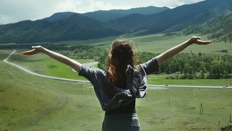 A beautiful girl reaching the top of the mountain, raises her hands and looks at the landscape.