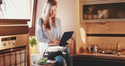 Young Woman Drinking Coffee Using Digital Tablet at Sunny Kitchen. SLOW MOTION 120 fps, 4K DCi. Relaxed Girl enjoying cozy time at sunlit home, eating breakfast or lunch, communicating online.