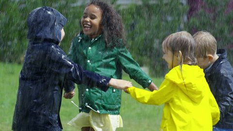 Slow motion with tilt down of happy little boys and girls in rubber boots and raincoats holding hands and running and jumping up in circle on green grass during heavy rain