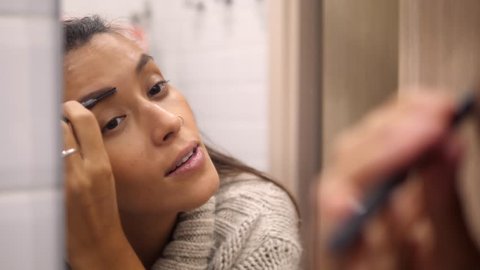 Attractive Young Mixed Race Woman in Winter Sweater Looks into Mirror and Paint Eyebrows