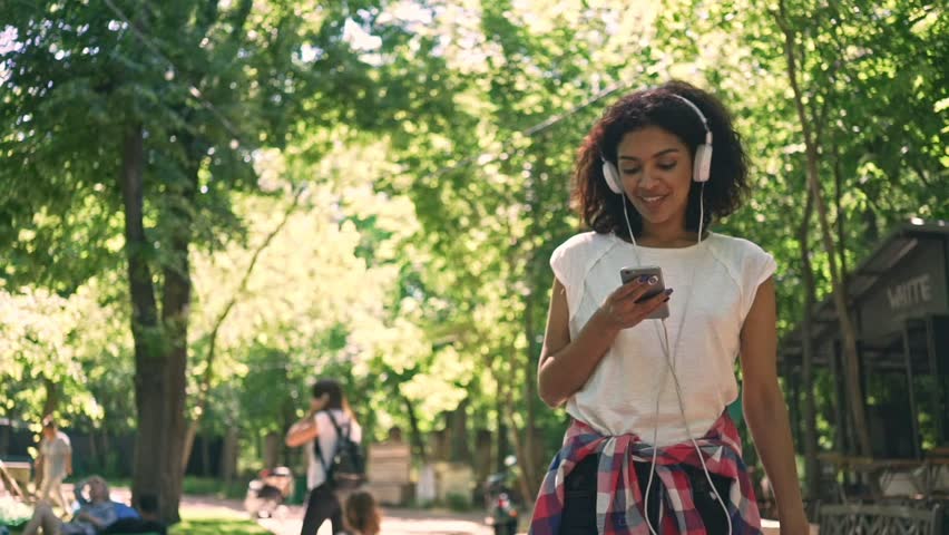 Smiling young african lady walking at the park outdoors chatting by phone listening music
