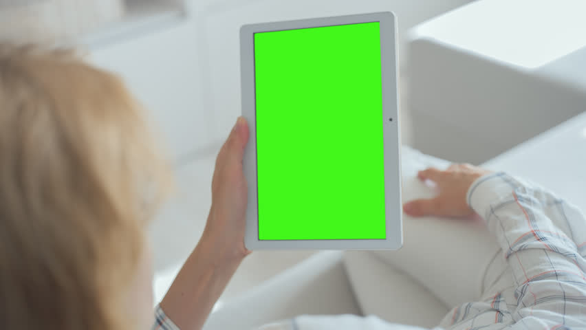 Young Woman in white jeans laying on couch holds Tablet PC with pre-keyed green screen. Perfect for screen compositing