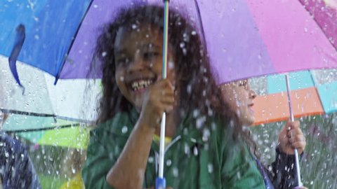 Medium shot with slow motion of happy little African-American girl with curly hair and her preschool friends holding colorful umbrellas and jumping in heavy rain