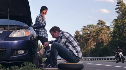 handsome caucasian man changing a tire on the side of the road. Young woman wearing casual clothes has phone conversation on smartphone . Female looking on the guy and smiling.