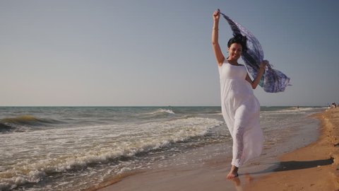Gypsy young brunette girl wearing white maxi long dress running near sea or ocean windy beach with mandala silk scarf in hands.Bohemian style. Boho lifestyle.Slow motion.Film tonned