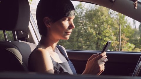 Adult woman sitting on the driver’s seat in car holding smartphone messaging or chatting with friend. Brunette with short hair using internet for send email or texting message.