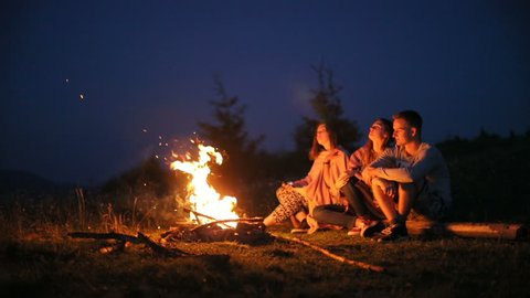 Young people sitting fire camp looking sky night outside nature pointing finger stars romantic having rest friends girls boy woman man three persons bonfire camping silence calm summer holidays warm స్టాక్ వీడియో