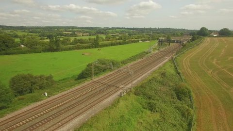 Aerial shot of a small local passenger train passing through the UK countryside in front of the camera as it tracks back. The train briefly passes behind a tree. Ungraded.