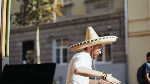 Mexican party concept - portrait of young attractive bearded man with glasses in casual wear with mexican sombrero dancing at the city urban old architecture street