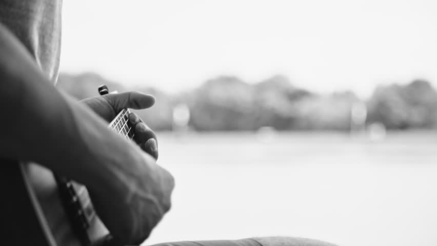 Black and white crop shot of man holding and playing on little acoustic ukulele guitar sitting in the outdoor park