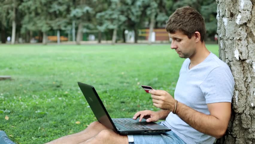 A man with a laptop in the park uses a bank credit card.