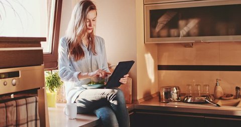 Young Woman Drinking Coffee Using Digital Tablet at Sunny Kitchen. SLOW MOTION 120 fps, 4K DCi. Relaxed Girl enjoying cozy time at sunlit home, eating breakfast or lunch, communicating online.