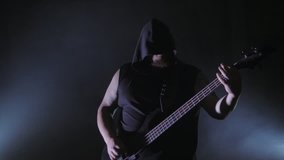 A brutal man in a hood playing bass guitar. Music video punk, heavy metal or rock group.