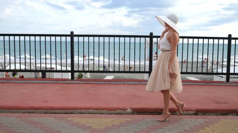 An elegant woman in a hat and a white air dress strolls along the waterfront. General shooting. 4k, slow-motion