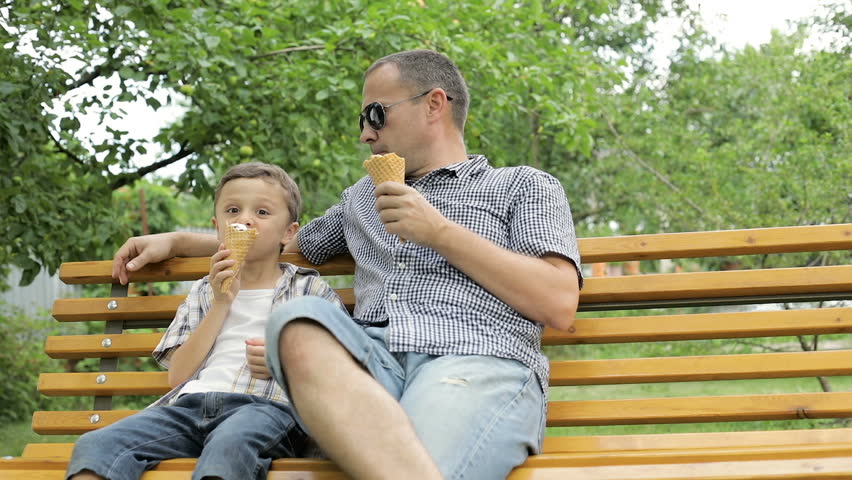 Father and son sitting on the bench in the park at the day time. They eating ice cream. Concept of friendly family.