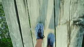 POV video: The feet of a woman are walking along a narrow wooden bridge over a mountain river. Danger and adventure