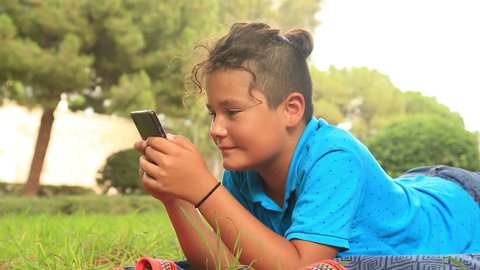 Preteen boy sitting on lawn with smart phone texting message or playing game at home. Technology, internet communication and people concept
