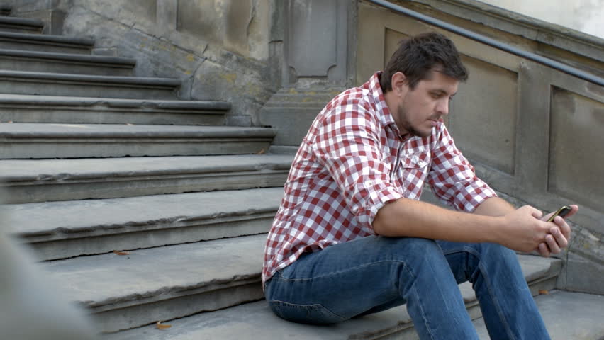 A young sad man is sitting on the old stairs and using app on the smartphone Royalty-Free Stock Footage #30312484