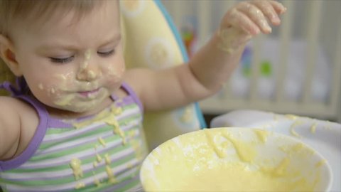 A small child alone eats baby food with a spoon from a saucer spilling into porridge