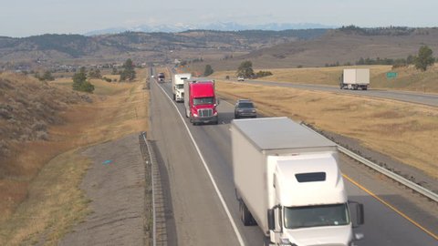 AERIAL, SLOW MOTION, CLOSE UP: Scenic highway on grass prairie with Rocky Mountains Range in the background. Cars traveling road trip, freight semi trucks transporting goods, SUVs & pickups driving