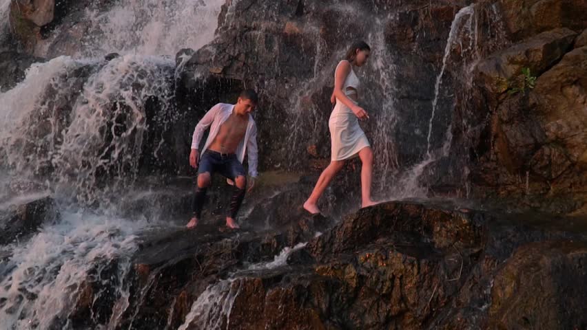 A guy and a girl in a white dress neatly make their way among the rocks under a waterfall during sunset