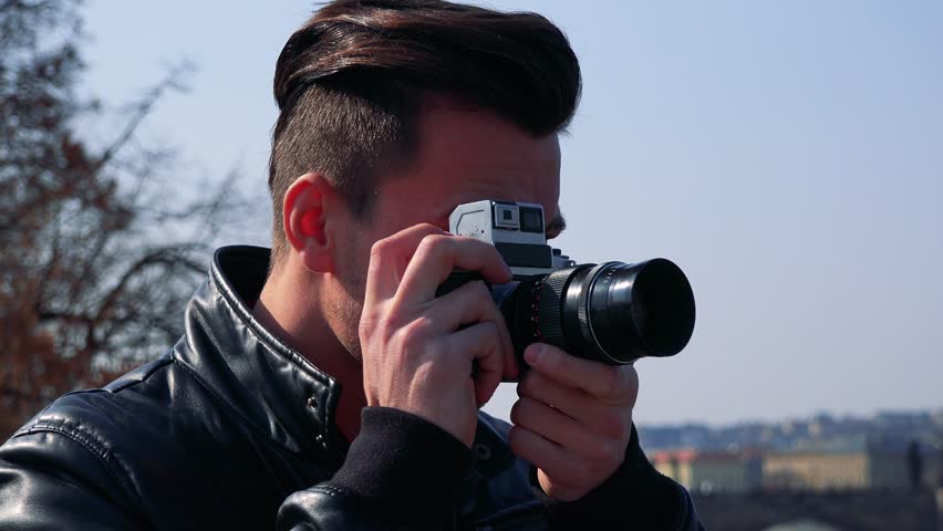 A young handsome man takes photos with a camera - face closeup - the blue sky and a townscape in the blurry background