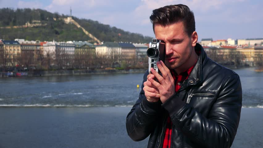 A young handsome man shoots a video with a camera - closeup - a river and a quaint townscape in the blurry background