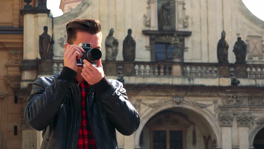 A young handsome man takes photos with a camera - a quaint building in the blurry background