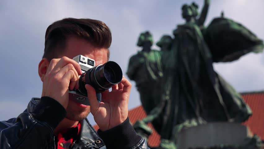 A young handsome man takes photos with a camera - closeup from below - a statue in the blurry background