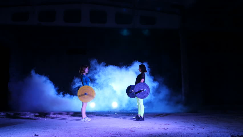 Two athletic girls, athletes, raise the barbell, do sit-ups with the barbell. At night, in the light of searchlights, in light smoke, fog, in an old abandoned hangar, the building.