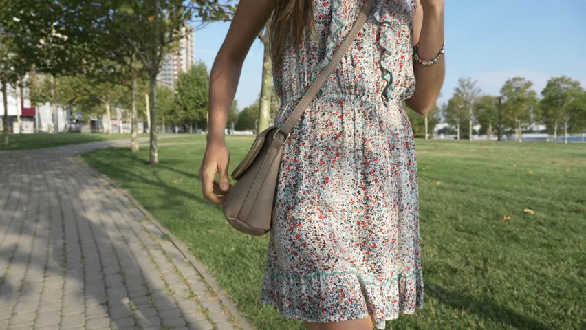 Young happy woman in dress pulls out from the handbag smart phone and starts talk while walking in city park, 4K slow motion steadicam shot