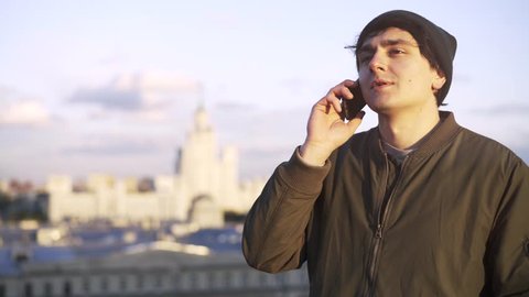 Serious young man wearing a khaki jacket and a beanie is talking on his smartphone while standing on a roof of a building. Locked down real time medium shot