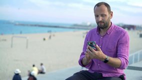 Happy man chatting on smartphone sitting near beach in city
