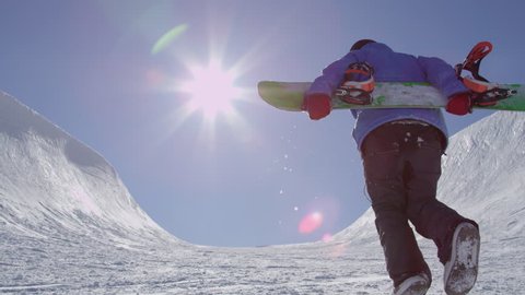 SLOW MOTION: Young pro snowboarder walking uphill in the halfpipe snow park carrying his board on a beautiful sunny day in snowy winter
