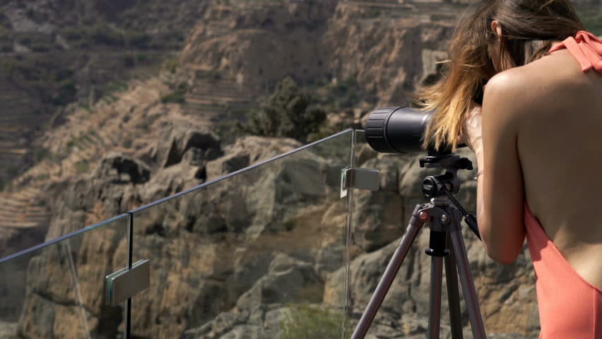 Woman admire canyon with telescope standing on terrace in mountains
