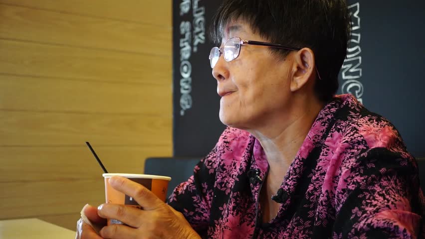 old senior Asian woman wear glasses, holding hot coffee cup looking around alone in the restaurant