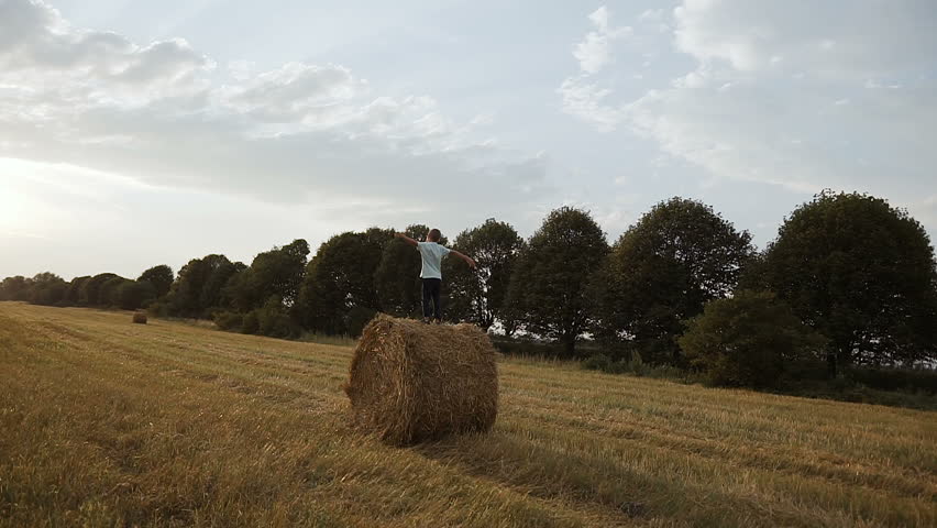 In a field on a haystack there is a boy and looks at the sunset slow motion