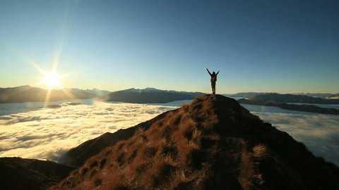 Young woman hiker stands alone on mountain summit above the clouds at sunset, arms outstretched as a sign of achievement and success.
Standing on top of the world, individuality on a mountain top 