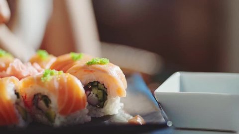 Woman Hand With Chopsticks Dipping Sushi Roll into Soy Sauce. 4K Close Up. Girl eating sushi with salmon on outdoor terrace of Japanese restaurant.