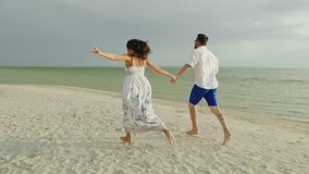 Young man and woman hold hands, run barefoot along the beach. Happy Holidays and Honeymoon. 4K slow motion video