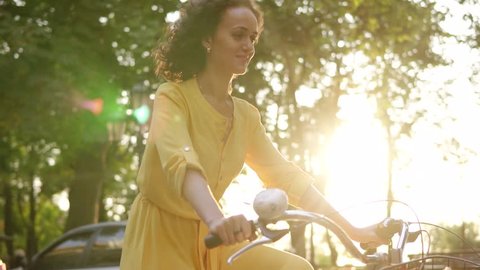 Lens flare: smiling happy woman in long yellow dress is riding a city bicycle with a basket and flowers in the park with green trees around during the dawn. Slowmotion shot