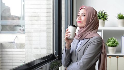 attractive beauty lady muslim business worker holding coffee mug standing in office and looking at window outside daydreaming relaxing.