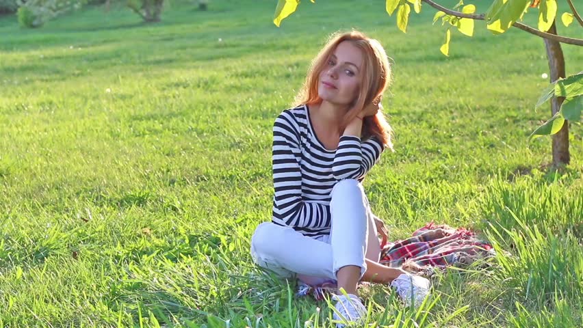 Young beautiful woman enjoying summer sunset sun light in the park, sitting on the grass, sensually smiling at camera, touching long blonde hair