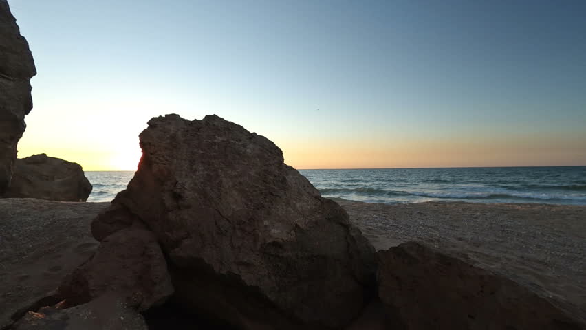 Coastal twilight granduer: rugged rock formations create dramatic silhouette along serene shoreline during sundown. Weathered stones occupy foreground with textured surfaces in earthy tones, contrasting against soft golden sands stretching toward water