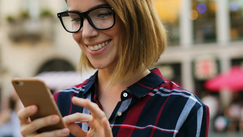 Caucasian woman with glasses using smart phone, typing something during walking in the city street. Portrait shot.