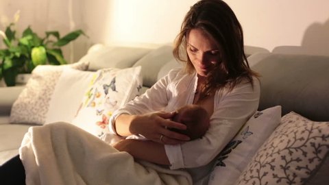 Young beautiful mother, breastfeeding her newborn baby boy at night, dim light. Mom breastfeeding infant