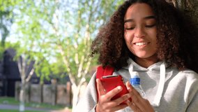 4K video clip of beautiful happy mixed race African American girl teenager leaning against a tree with a red backpack drinking from a water bottle and using a cell phone app or text messaging 