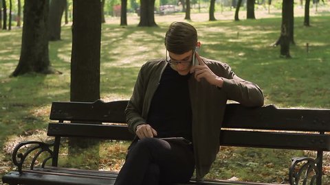 A young man with glasses sitting on a bench in the park works in an application on an electronic tablet