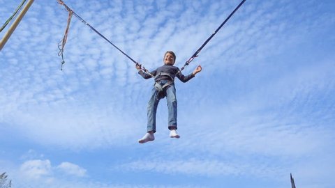 Young boy jumping on bungee trampoline against blue sky, slow motion