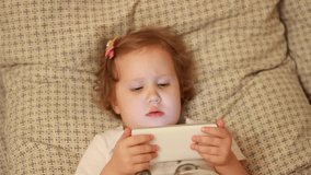Child Looks cartoons and plays downloaded application on a smart phone close-up. A girl lies in bed and looks at the phone screen.
